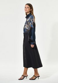 Pitkähihaiset kukalliset paita tummansinisessä ja mustassa, yhdistetty mustan midi-hameen kanssa, jossa on etupainikkeet ja sivutaskut. Yhdistetty mustien korkokenkien kanssa.