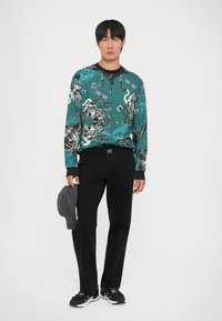 Camicia a maniche lunghe color turchese con grafiche nere, pantaloni neri con cuciture bianche e sneakers nere. Tieni in mano un cappellino grigio.