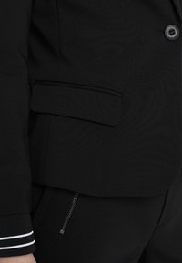 Zwarte blazer met een gemêleerde stof, een enkele knoopsluiting, zijzakken en contrasterende witte strepen bij de manchetten.