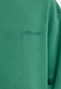 Zielony materiał z bliska z subtelnym haftowanym logo "element" na wysokości klatki piersiowej ubrania.