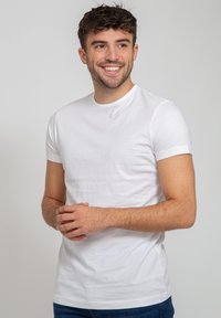 Weißes Baumwoll-T-Shirt mit kurzen Ärmeln, geripptem Rundhalsausschnitt und taillierter Passform, getragen von einer Person mit gefalteten Händen vor der Brust.