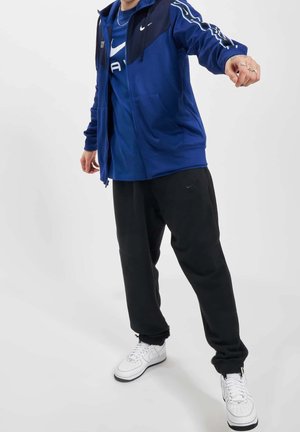 Veste zippée bleue avec manches sombres, pantalon de survêtement noir et baskets blanches. Présente des logos Nike et une coupe confortable.
