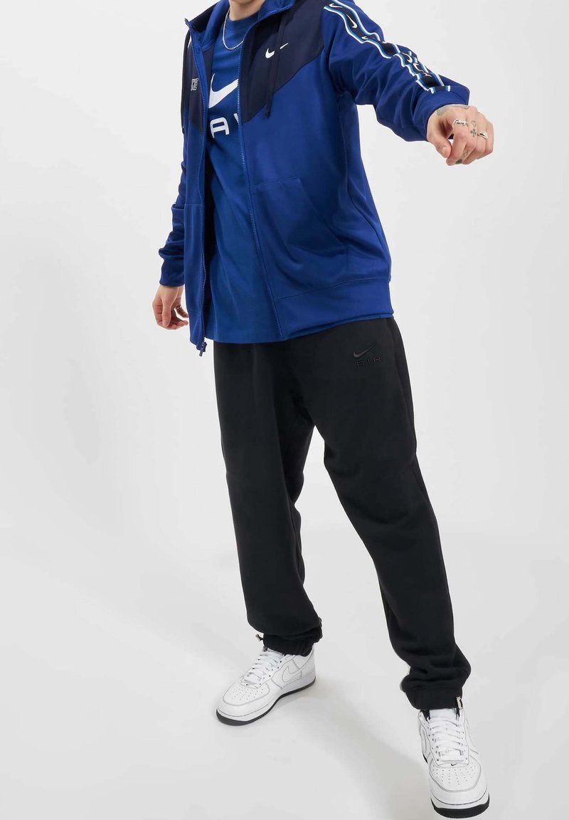 Blå zip-jacka med mörka ärmar, svarta sweatpants och vita sneakers. Har Nike-logotyper och en bekväm passform.