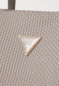 Sac à main beige tissé texturé avec une plaque de logo triangulaire dorée et blanche Guess U.S.A. sur le devant.