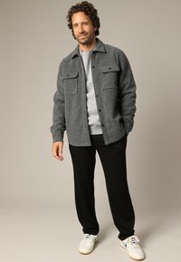 Grijze wollen blend shirtjack met twee borstzakken, knoopsluiting, gedragen over een lichtgrijze gebreide trui en zwarte broeken, gecombineerd met sneakers.