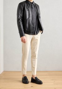 Svart läderjacka med hög krage, randig skjorta, krämfärgade slim-fit-byxor och svarta loafers med strukturerade sulor.