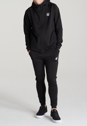 Schwarzer Kapuzenpullover mit Reißverschluss und Logo auf der Brust, kombiniert mit schwarzen, schmal zulaufenden Jogginghosen. Beide Kleidungsstücke haben eine glatte Stoffstruktur. Schwarze Sneaker vervollständigen das Outfit.