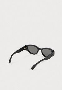 Μαύρα γυαλιά ηλίου με σχήμα γάτας, πλαστικό σκελετό και γκρίζους φακούς. Υπογραφή λεπτομέρειες μεταλλικών στοιχείων και λογότυπο στο πλάι.