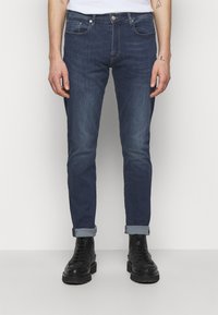 Man med mörkblå upprullade slim fit jeans och svarta snörkängor, stående mot en ljus enfärgad bakgrund.