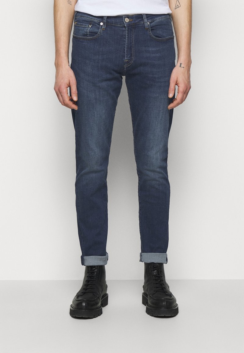 Man med mörkblå upprullade slim fit jeans och svarta snörkängor, stående mot en ljus enfärgad bakgrund.