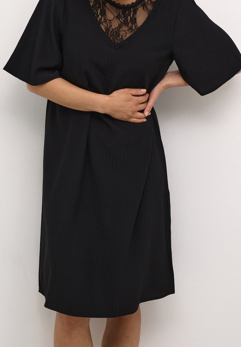 Femme portant une robe noire avec un décolleté en dentelle, tenant son ventre et le bas de son dos, debout devant un fond clair uni.