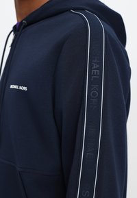 Felpa con cappuccio blu navy, caratterizzata da una zip frontale, strisce bianche decorative lungo le maniche e il logo "Michael Kors" in rilievo.