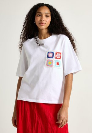 Ung kvinde med krøllet hår iført en hvid oversized T-shirt med farverig hæklet firkantet lap og en rød nederdel, stående mod en ensfarvet baggrund.