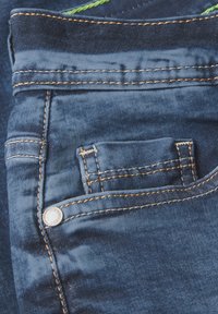 Pantalons en tissu denim avec une poche avant ornée de coutures oranges contrastantes et une petite poche décorative au-dessus. Couleur bleu foncé texturé.