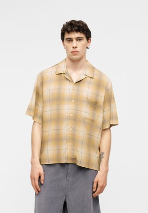 Mladić s kratkom kovrčavom kosom, odjeven u široku žutu kariranu košulju kratkih rukava i sive hlače, stoji ispred jednobojne pozadine.