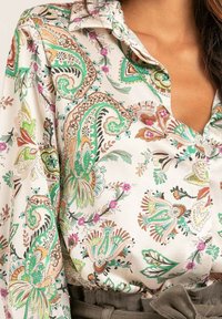 Blouse en soie de couleur claire, ornée d'un motif floral paisley dans des teintes de vert, brun et rose. Elle possède un col pointu et une fermeture à boutons.
