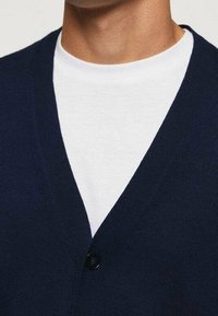 Cardigan bleu marine à col en V, présentant une texture côtelée et une fermeture à un bouton noir, porté sur une chemise blanche.