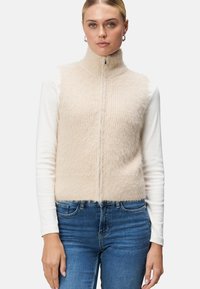 Gilet beige en peluche sans manches avec un col montant et une fermeture éclair à l'avant, superposé à un haut blanc à manches longues, associé à un jean bleu.