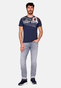 Mornarska majica s grafičnim tiskom in napisom; svetlo sivi jeans; temni čevlji; splošna casual obleka z udobnim krojem.