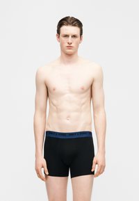 Νέος άνδρας με ανοιχτόχρωμο δέρμα και σκουρόχρωμα μαλλιά στέκεται μπροστά σε απλό φόντο, φορώντας μαύρα μποξεράκια με μπλε ζώνη μέσης που αναγράφει "ONLY & SONS".