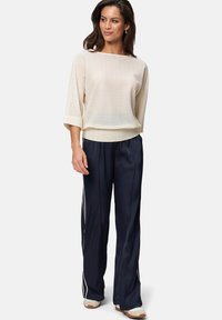 Haut tricot texturé beige clair avec une coupe ample et des manches trois-quarts, associé à un pantalon en satin bleu marine avec des rayures blanches sur les côtés.