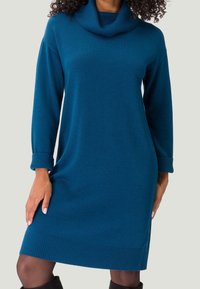 Abito lavorato a maglia verde blu con scollo a cratere, polsini e orlo a coste. Silhouette fluida, maniche lunghe, tessuto morbido, taglio a ginocchio.