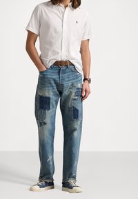 Vit kortärmad skjorta med knappar, blå slitna denimjeans med lappar, ljusblå sneakers med mörka detaljer. Brun bälte och armbandsur.