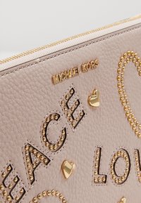 Pochette en cuir beige texturé avec le texte "PEACE" et "LOVE" en clous dorés, des cœurs dorés et le logo Michael Kors doré près de la fermeture éclair.