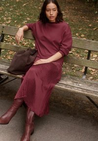 Burgundy tröja och plisserad kjol, brun mockaväska, knähöga bruna stövlar, sittande på en bänk i parken med höstlöv i bakgrunden.