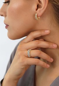 Woman wearing gold hoop earring and matching gold ring with blue and white gemstones, fingers gently touching her jawline.