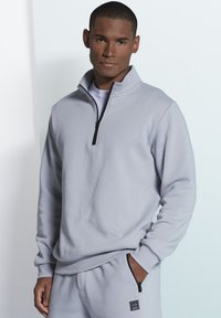 Gray fleece pullover with a zippered collar and long sleeves, featuring a textured surface and a small logo accent on the lower left.