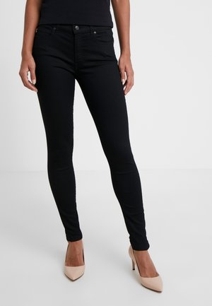 Femme portant un jean skinny noir et des escarpins couleur nude, debout devant un fond clair uni.