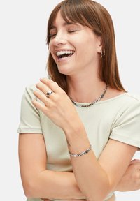 Silberne Kette mit kreisförmigen Akzenten, passender silberner Ring und Armband. Das Model trägt ein hellgrünes, geripptes Shirt und hat glattes braunes Haar.