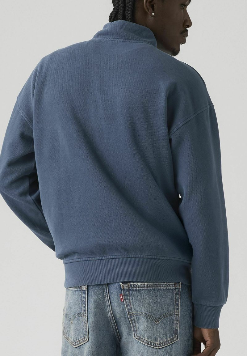 Hombre con cabello trenzado, vistiendo una sudadera azul y jeans de lavado claro, de perfil contra un fondo liso y claro.