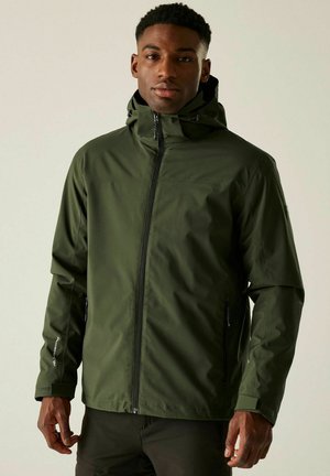 Jeune homme portant une veste d'extérieur à capuche vert olive avec fermeture éclair, poches zippées et poignets ajustables, se tenant devant un fond uni.