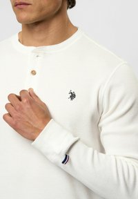 Mand iført en hvid, tekstureret langærmet skjorte med to træknapper og et lille sort broderet logo af en polo-spiller på brystet.