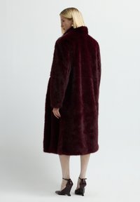 Nainen, joka pitää yllä pitkää tumman viininpunaisia tekoturkisvillapukua, valkoisia kuviollisia sukkahousuja ja tumman viininpunaisia korkokenkiä, seisoo tasaisessa taustassa.