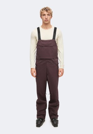 Jeune homme portant un pantalon salopette marron foncé avec des bretelles noires, une chemise beige à manches longues et des chaussures noires, debout devant un fond uni.