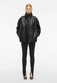 Veste en cuir noir avec grandes poches, col montant et fermeture éclair, portée sur un col roulé marron et associée à un pantalon noir à jambes fendues.