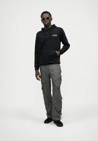 Zwarte hoodie met een voorkantzak en logo, gecombineerd met grijze cargobroek met meerdere zakken en een losse pasvorm. Zwarte sneakers en een zonnebril.