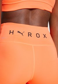 Narančaste atletske kratke hlače s visokim strukom, koje sadrže crni natpis "HROX" i Puma logo, napravljene su od materijala koji upija vlagu - DryCell.