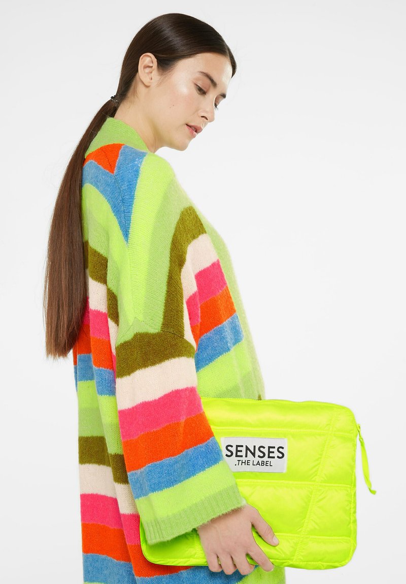 Gestreifter, übergroßer Strickpullover in Grün, Pink, Orange und Blau; hält eine neongrüne gepolsterte Laptophülle mit Logo.