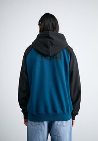 Persona con lunghi capelli scuri che indossa un felpa North Face blu e nera e jeans chiari, voltato di spalle su uno sfondo neutro.