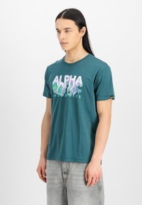 Tyrkysové bavlněné tričko s vlnitým grafickým motivem "ALPHA" v světle fialové a zelené. Krátké rukávy a volný střih. Ozdobené logem.