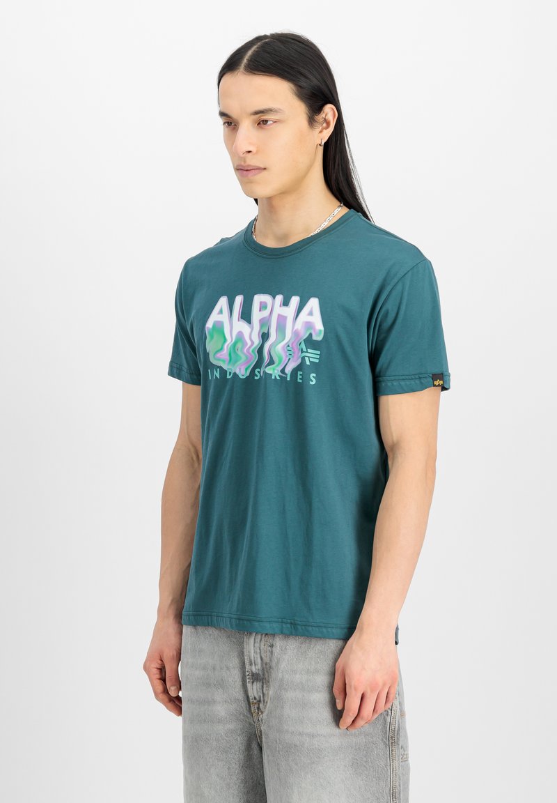 Turkizna bombažna majica z valovitim grafičnim napisom "ALPHA" v svetlo vijolični in zeleni barvi. Kratkimi rokavi in sproščenim krojem. Poudarjena z oznako logotipa.