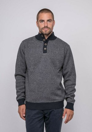 Homme aux cheveux courts portant une barbe, vêtu d'un pull-over à boutons gris rayé et d'un pantalon foncé, debout devant un arrière-plan uni.