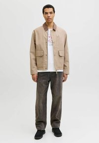 Smėlinis lengvas švarkas su rudomis aksomo apykaklėmis, dviem priekiniais kišenėmis, dėvimas ant balto marškinėlių su grafika ir anglies spalvos džinsais, derinamas su juodais batais.