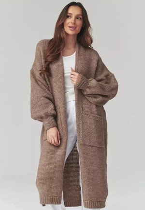 Kobieta w długim, oversize'owym, brązowym dzianinowym kardiganie z dużymi rękawami i przednimi kieszeniami, w białej bluzce i białych spodniach, pozująca na jednolitym tle.