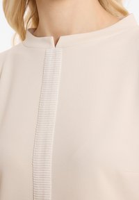 Helles beige Kleidungsstück mit einem hohen Kragen und einer vertikalen Rippstruktur in der Mitte, aus weichem Stoff gefertigt. Verfügt über ein minimalistisches Design.