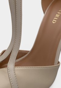 Sandale à talons hauts beige avec finition vernie brillante, dotée de deux fines lanières et d'une semelle intérieure lisse. Rehaussée de détails soignés en couture.